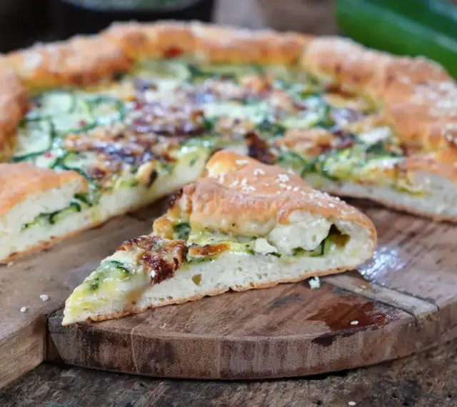 Leckeres Zucchini Pizza Rezept auf einem Holzbrett, mit einem Stück abgetrennt.