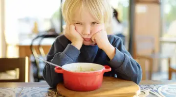 Ein Junge mit blonden Haaren stützt seinen Kopf auf die Hände und schaut lustlos auf eine Schüssel Porridge. Er isst nicht, obwohl er 10 Jahre alt ist.