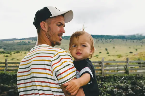 Ein Mann mit Bart und Kappe trägt ein Kind. Die Eigenschaften eines Papas zeigen sich in seiner liebevollen Umarmung.