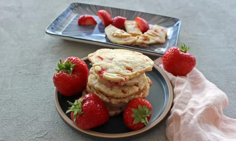 Ein Stapel Erdbeer-Pancakes mit frischen Erdbeeren auf einem Teller. Ein weiterer Teller im Hintergrund zeigt geschnittene Erdbeeren und Pancakes mit Erdnussbutter.