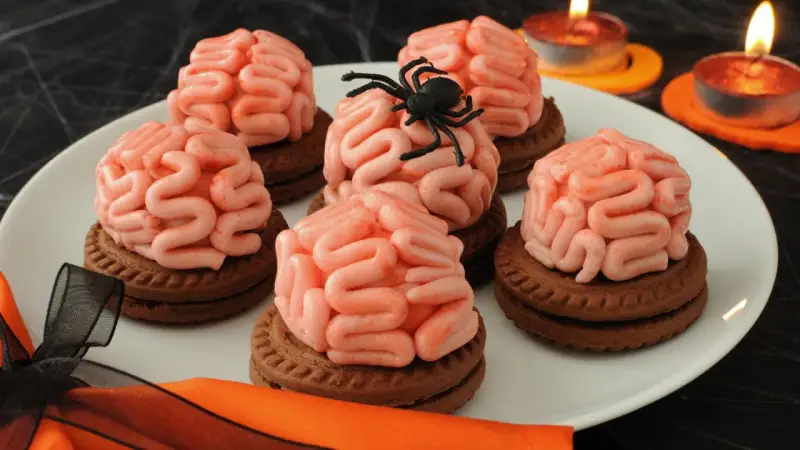 Gruselige Halloween-Nachspeise: Kekse mit Gehirn-förmiger rosa Glasur, dekoriert mit einer Spinne. Kerzen leuchten im Hintergrund.