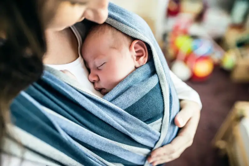 Ein Baby schläft friedlich in einem blauen Tragetuch. Der sanfte Tagesrhythmus des Babys wird durch die Nähe zur Mutter unterstützt.